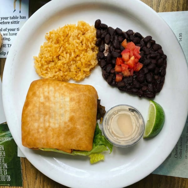 A golden-brown burrito, rice, black beans with diced tomatoes, sauce, and a lime wedge on a white plate.