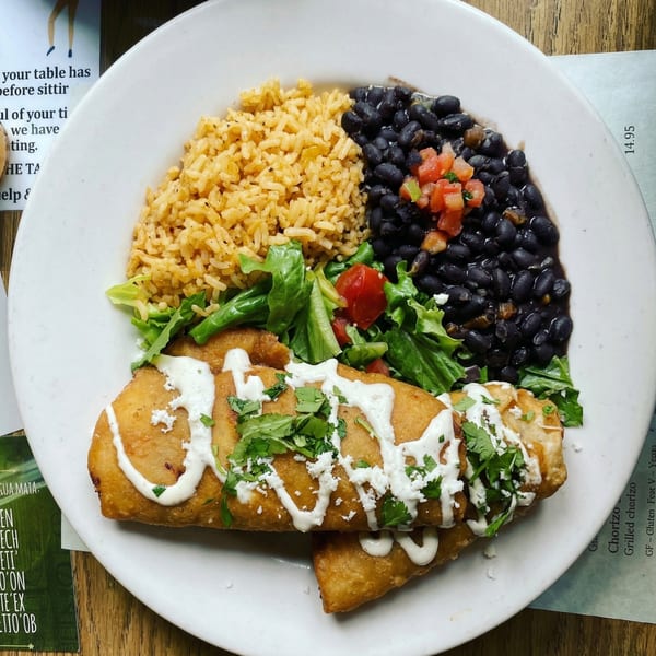 Two golden-brown fried burritos topped with crema, cilantro, and cotija cheese, served with rice, black beans, and a side salad.